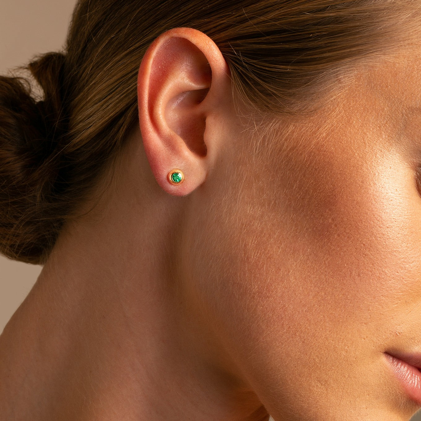 Close up on model 14K Yellow Gold Domed Bezel Set Emerald Stud Earrings. .20tcw