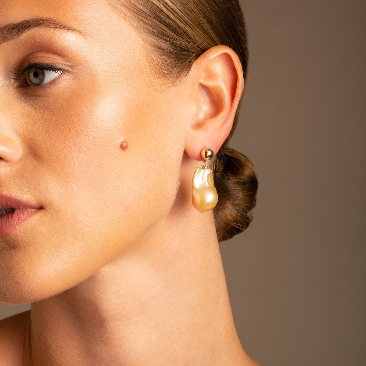 Featuring a sleek 7.5mm hollow 14k Yellow Gold ball with a glossy finish, this Double Bubble Design is adorned with a captivating golden-hued baroque pearl drop . On model photo